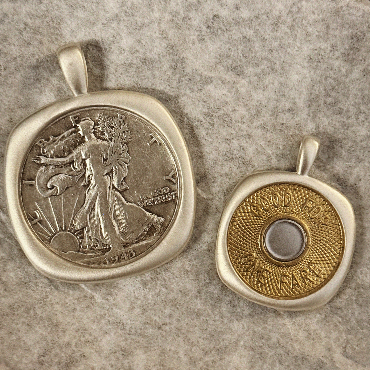 Two silver-toned coin pendants displayed on textured gray paper. The larger pendant on the left is set with a silver-colored coin, while the smaller pendant on the right features a gold-toned coin with a silver-colored circular center.
