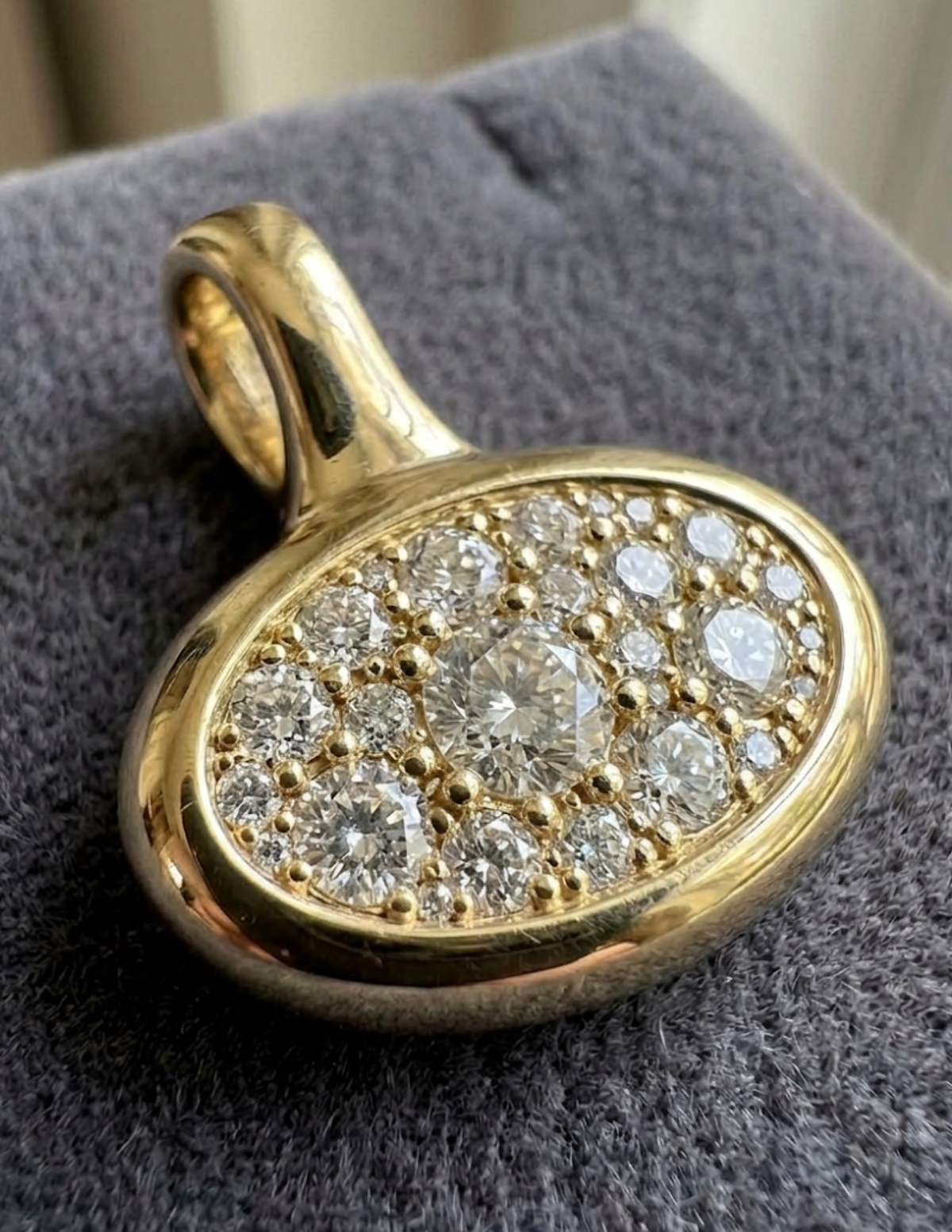 A gold oval pendant with a pavé setting of clear round gemstones, resting on a textured gray fabric surface.