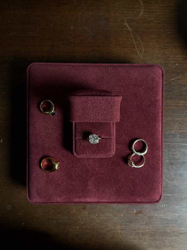 A top-down view of several rings displayed on a square burgundy velvet jewelry box placed on a dark wooden table.