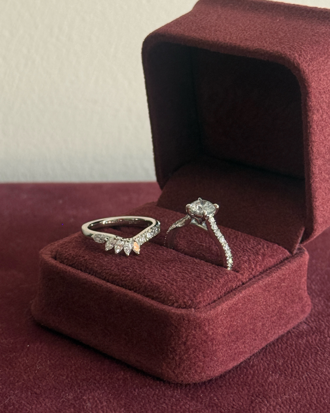 Two silver rings, one a band with small diamond accents and the other an engagement ring with a round center stone, displayed in an open dark red velvet ring box.