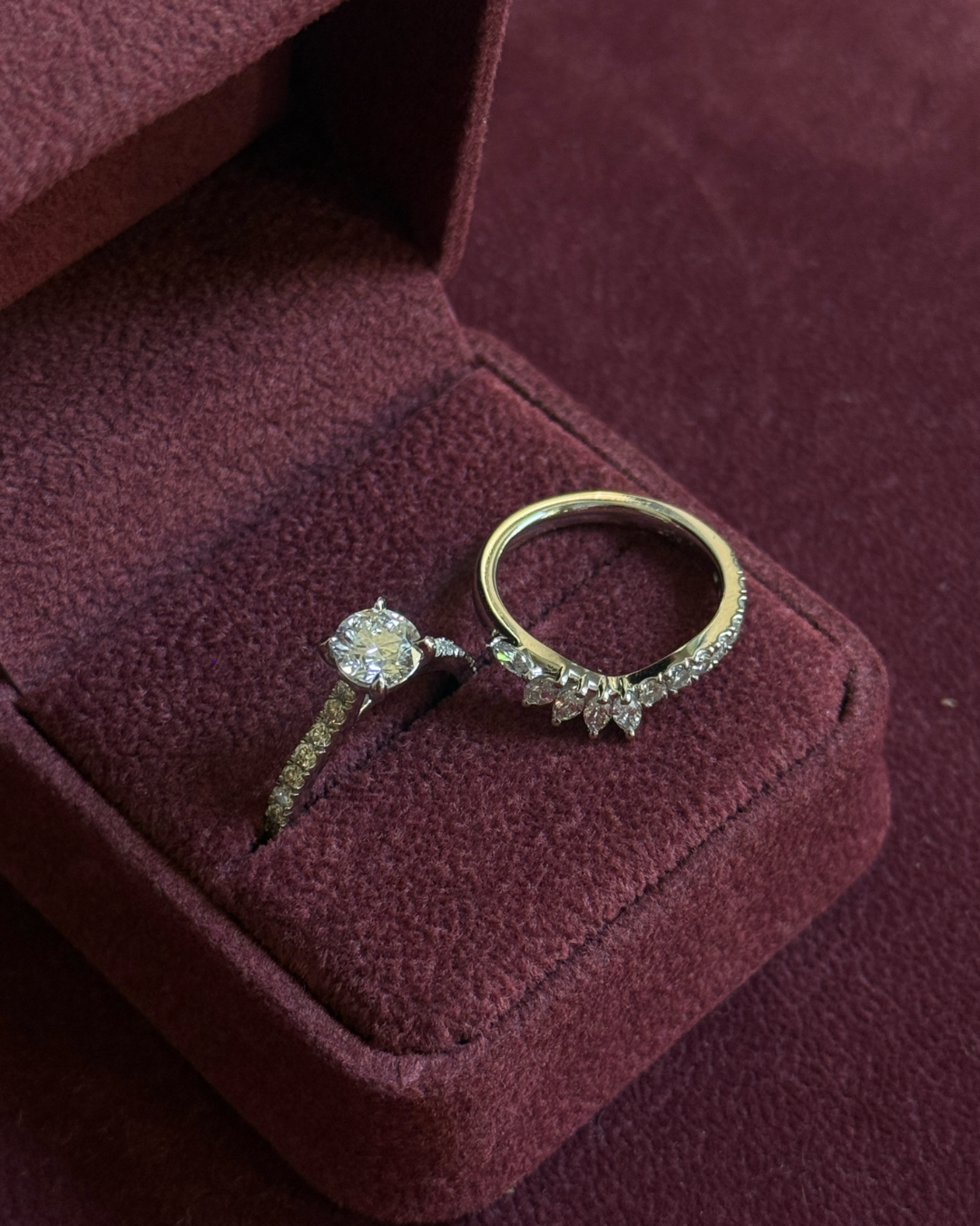 Two rings displayed in an open burgundy velvet jewelry box. One ring features a round clear gemstone on a gold-toned band, and the other is a yellow gold band with a row of small clear stones and a leaf-like centerpiece.