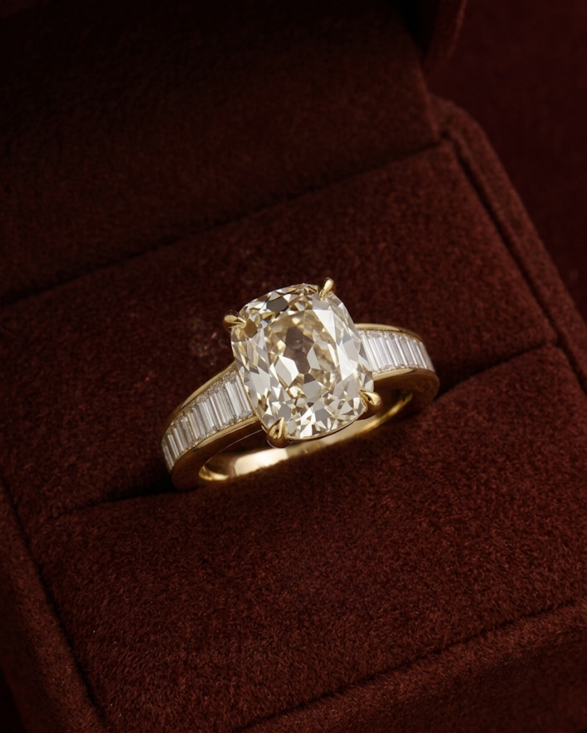 An oval-cut diamond ring with a yellow gold band featuring baguette diamonds, displayed inside a dark brown velvet ring box.