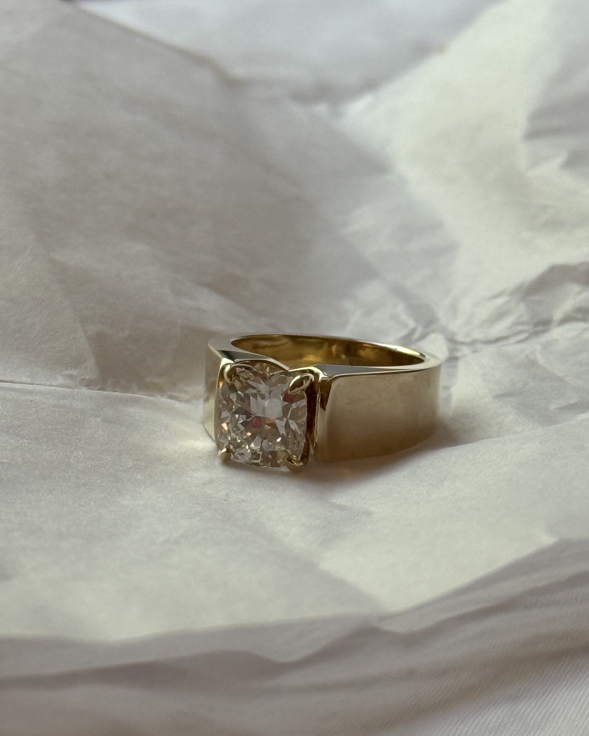 A yellow gold ring with a large cushion-cut clear gemstone in a four-prong setting, resting on crumpled white paper.