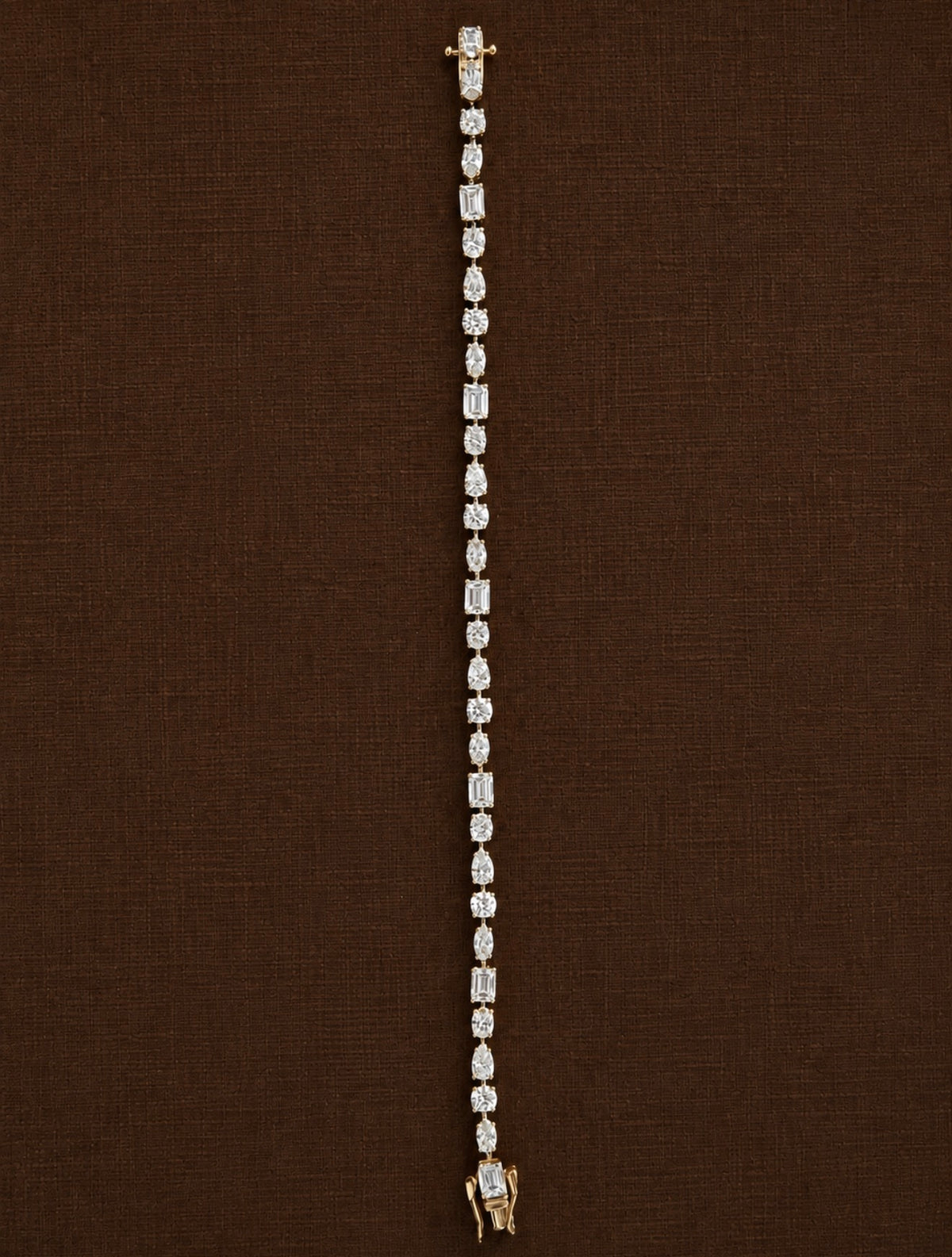 A delicate yellow gold tennis bracelet with a continuous line of round-cut clear gemstones, displayed vertically against a textured dark brown background.