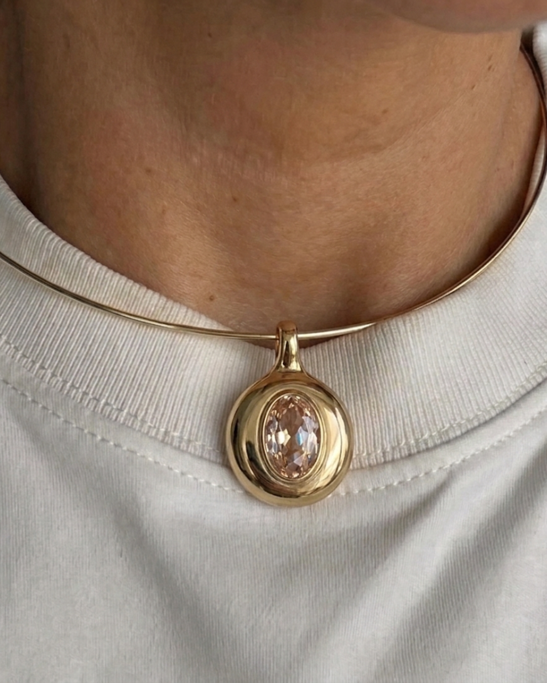 A close-up of a person wearing a gold bangle necklace with a round bezel-set peach-colored gemstone pendant, styled with a white crewneck top.