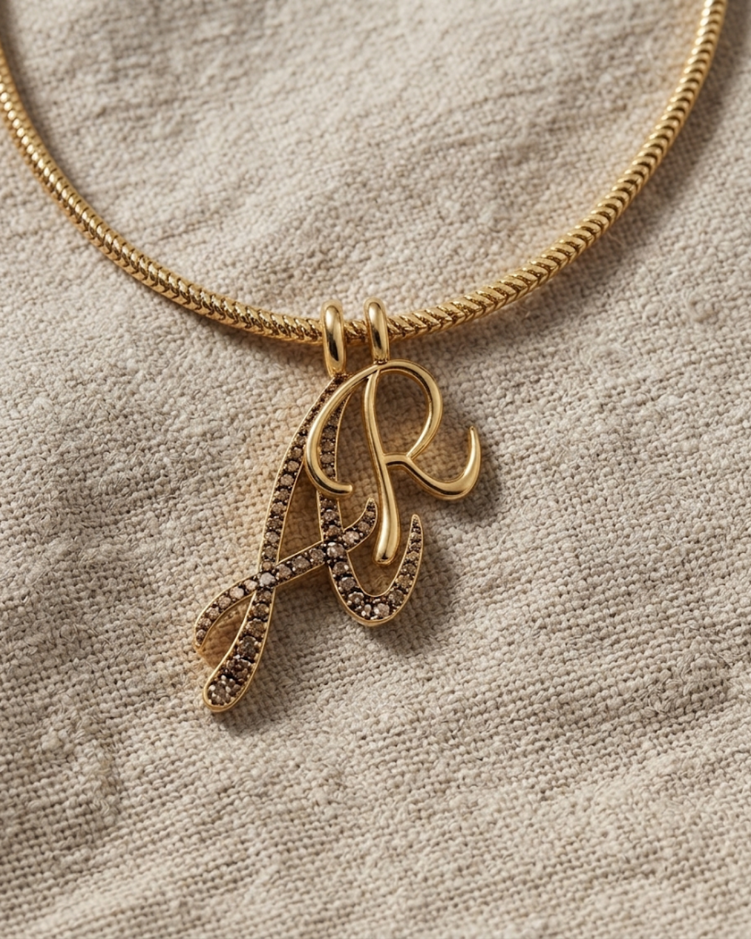 A gold chain necklace with a monogram pendant featuring the letters 'A' and 'R' in polished gold and pavé-set clear stones, displayed on a textured beige linen fabric.
